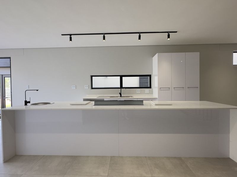 Modern, minimalist kitchen featuring a large central island with stone countertops, sleek white cabinetry, and contemporary track lighting in an open-plan layout.