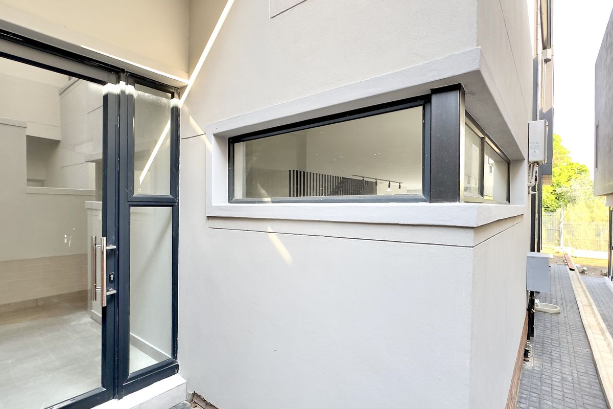 A modern exterior view of the townhouse featuring clean plastered walls, dark aluminium window frames, and a paved side walkway.