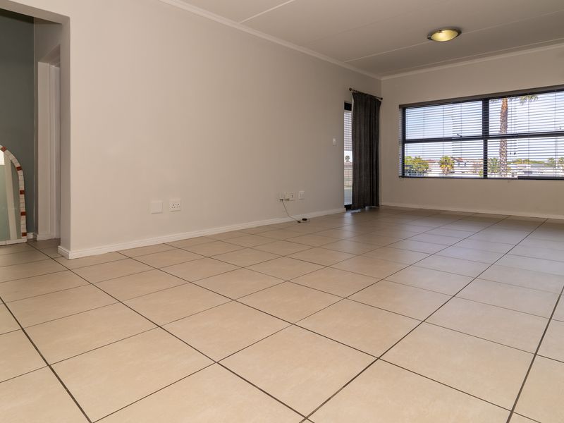 A spacious, empty living area featuring neutral tiled flooring, a large window with blinds, and a decorative archway leading to an adjacent space.