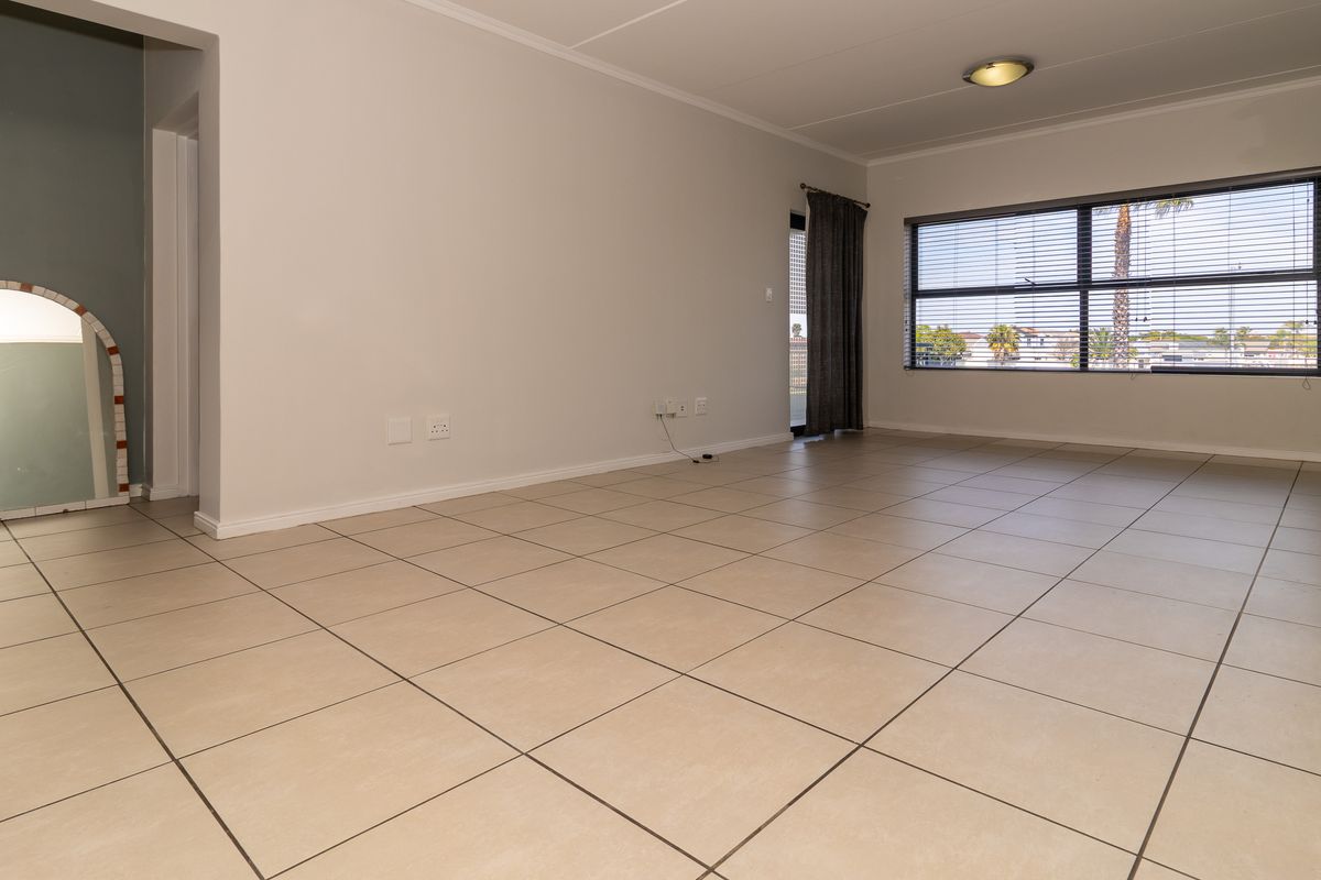A spacious, empty living area featuring neutral tiled flooring, a large window with blinds, and a decorative archway leading to an adjacent space.