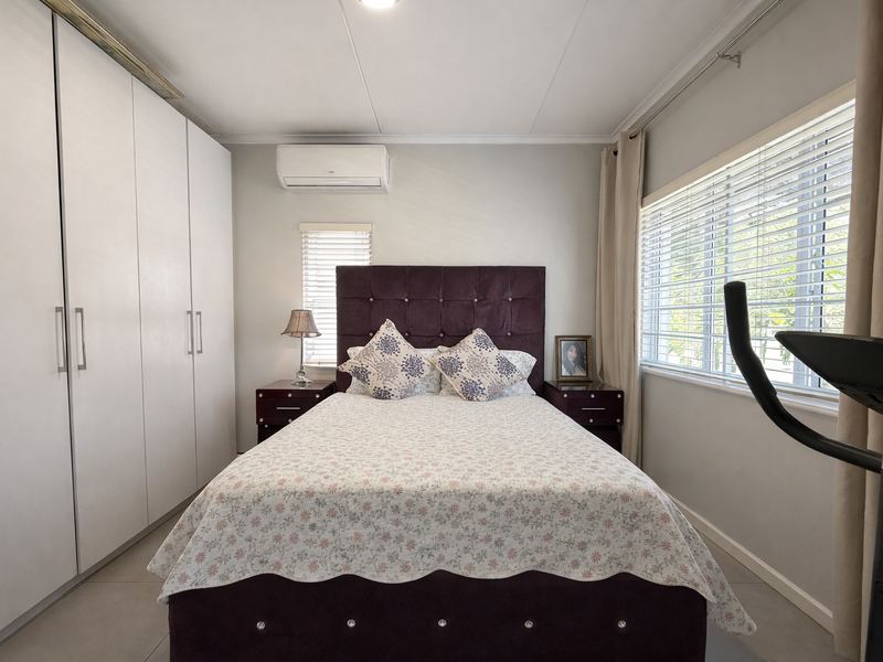 A well-maintained bedroom featuring built-in cupboards, tiled flooring, an air conditioning unit, and large windows with venetian blinds for natural light.