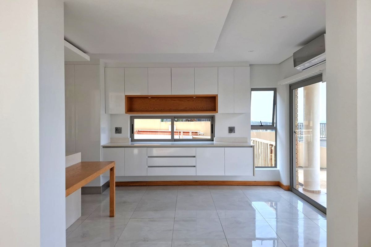 Modern, minimalist kitchen featuring sleek white cabinetry, wood accents, stone countertops, and large sliding glass doors leading to an outdoor area.