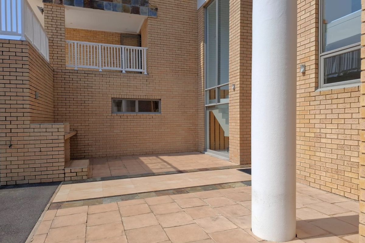 A spacious rear patio area featuring a built-in braai, brick facade, and access to an upper-level balcony with white railings.