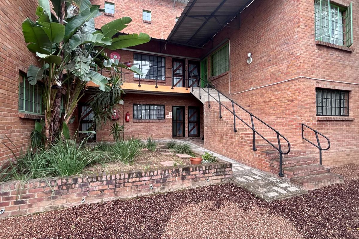 Exterior view of a brick apartment building featuring a staircase, security gates, and a small landscaped garden area with a brick retaining wall.