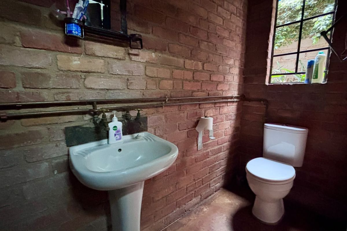 A rustic bathroom featuring exposed brick walls, a pedestal basin, and a toilet, with visible copper plumbing and a steel-framed window.