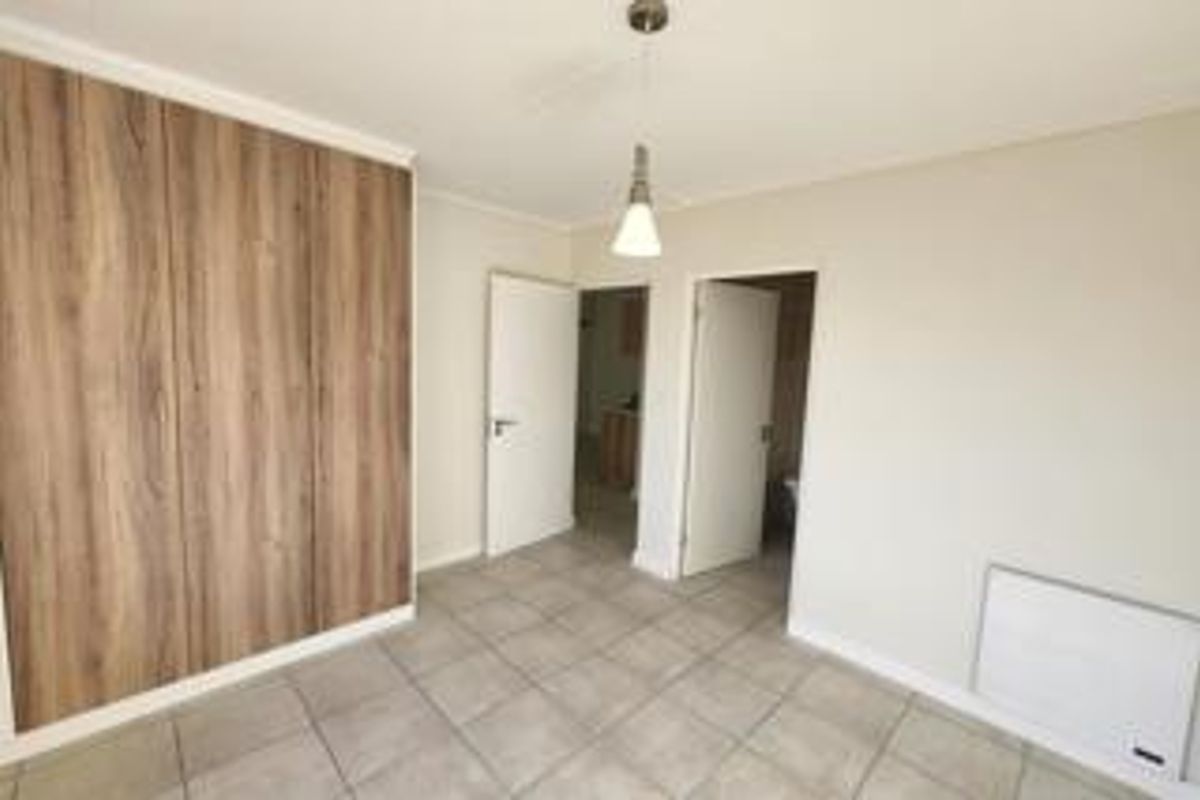 A bright, empty bedroom featuring modern wood-finish built-in cupboards, neutral tiled flooring, and a wall-mounted heater.