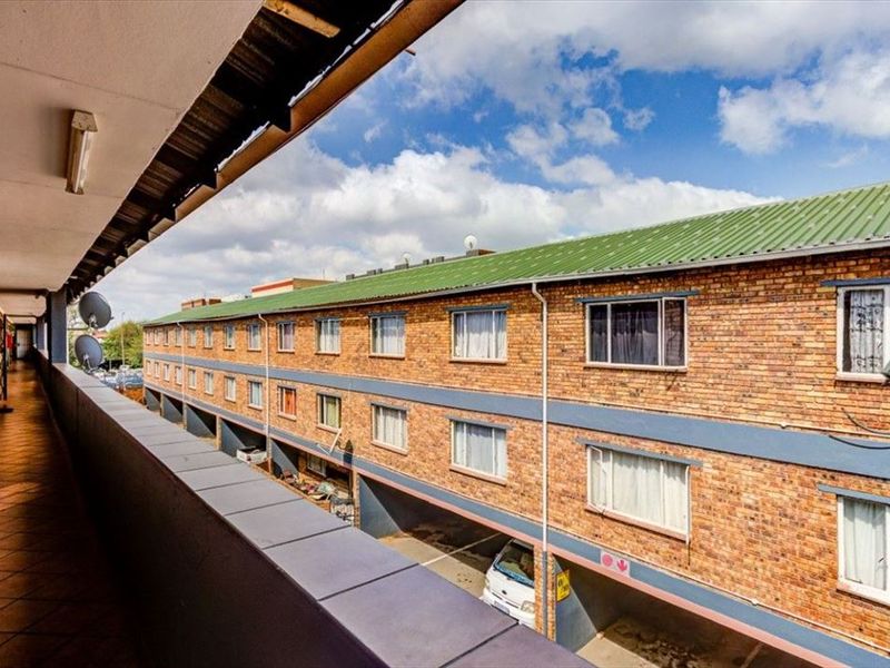 An exterior view from a covered balcony overlooking the apartment complex facade, featuring brick construction and a metal roof.