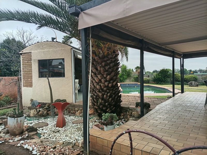 Covered patio area overlooking a swimming pool and garden, featuring paved flooring and an adjacent brick outbuilding.