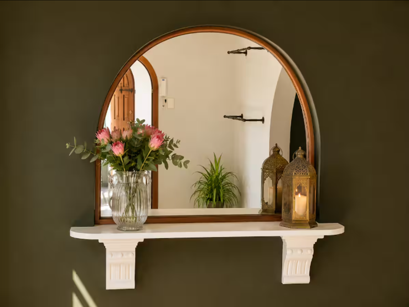 A decorative arched wall mirror mounted above a white shelf with ornate corbels, featuring an intercom system in the background.