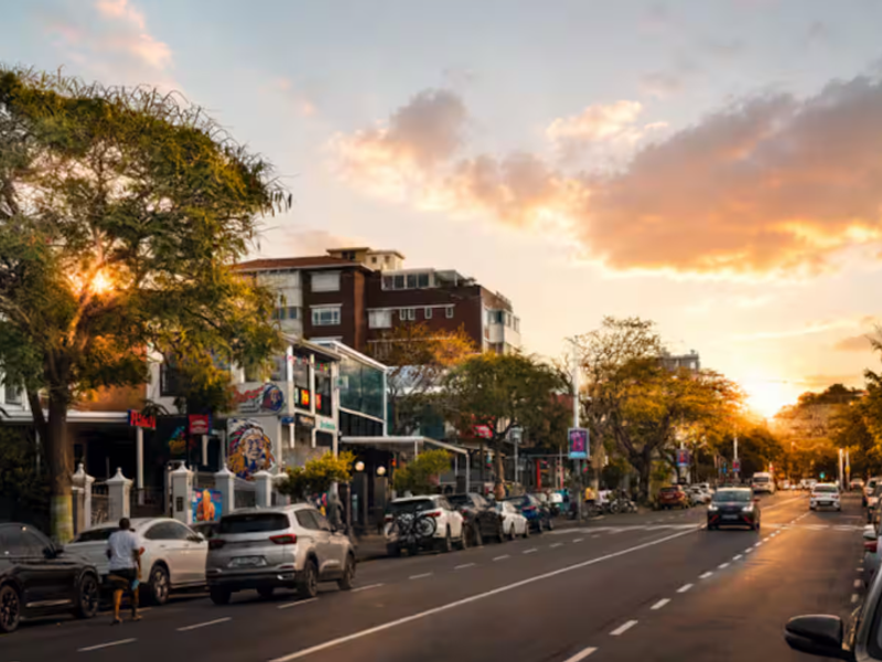A scenic street view of a neighbourhood road lined with buildings, trees, and parked vehicles during a sunset.