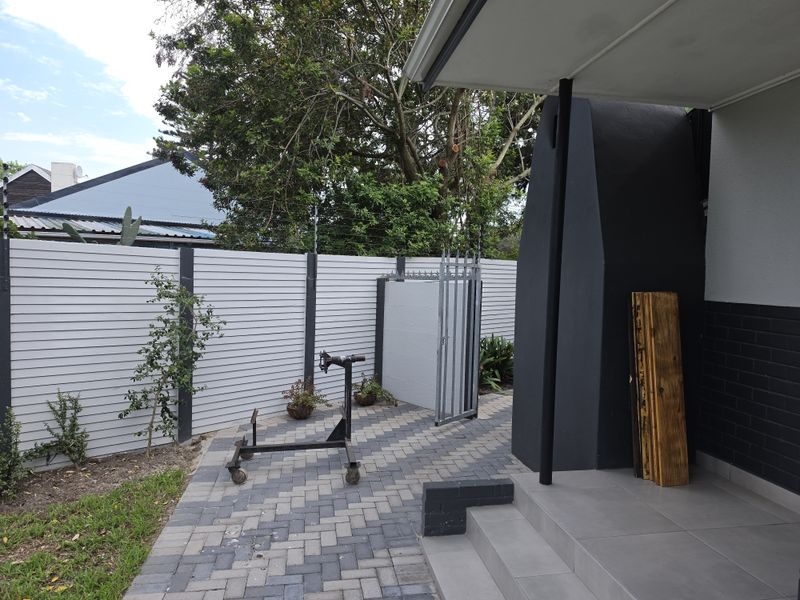 A covered patio area featuring a modern outdoor fireplace, paved flooring, and secure perimeter fencing with a gate.