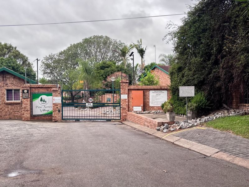 The entrance to Pine Acres Village, featuring a brick boundary wall, secure metal gate, and paved driveway access.