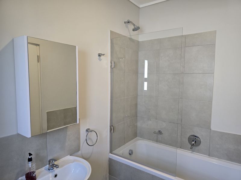 A clean, modern bathroom featuring a shower-over-bath combination with a glass screen, pedestal basin, and a built-in mirrored vanity cabinet.