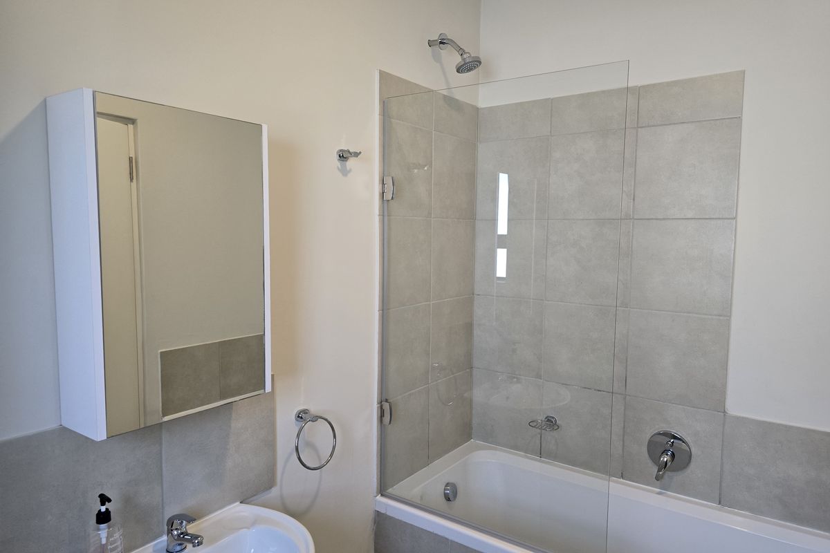 A clean, modern bathroom featuring a shower-over-bath combination with a glass screen, pedestal basin, and a built-in mirrored vanity cabinet.