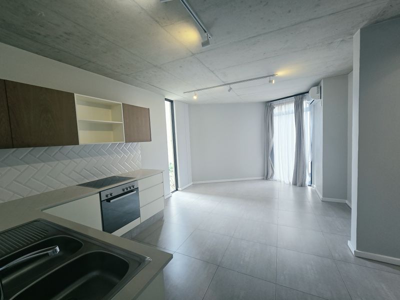 Modern open-plan living area featuring an exposed concrete ceiling, sleek kitchen with built-in appliances, herringbone backsplash, and neutral tiled flooring throughout.