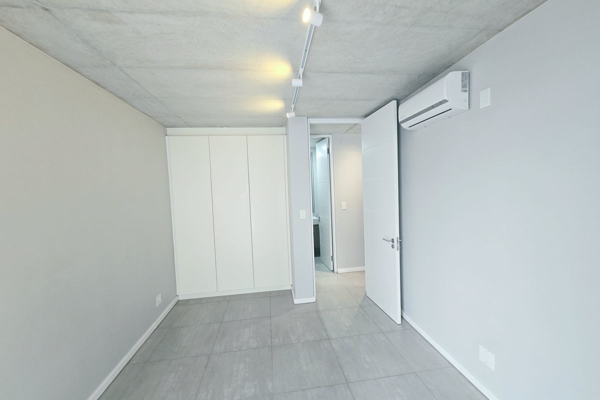 A modern, empty bedroom featuring an exposed concrete ceiling, built-in wardrobe, split-system air conditioning, and neutral tiled flooring with access to an en-suite bathroom.