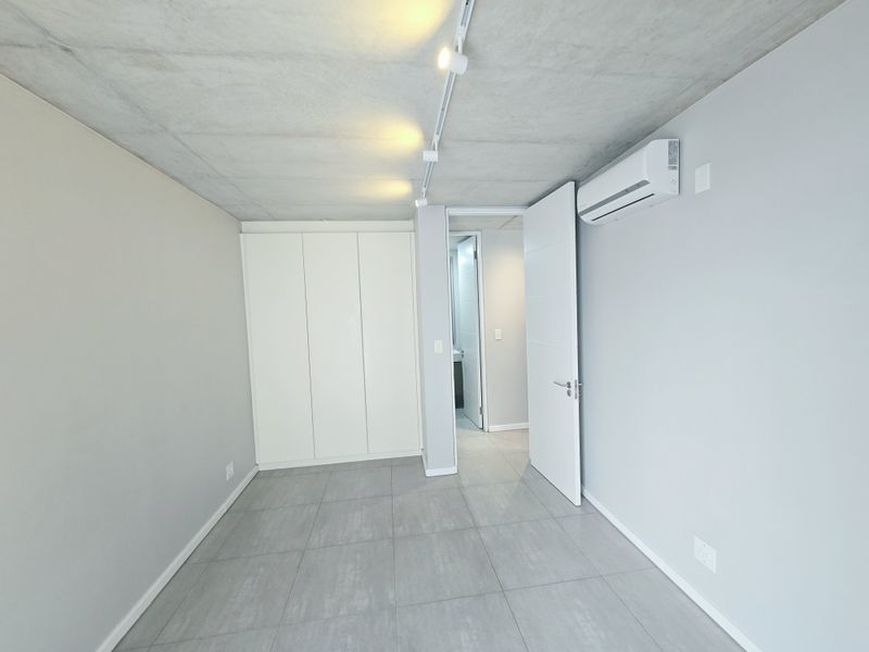 A modern, empty bedroom featuring an exposed concrete ceiling, built-in wardrobe, split-system air conditioning, and neutral tiled flooring with access to an en-suite bathroom.