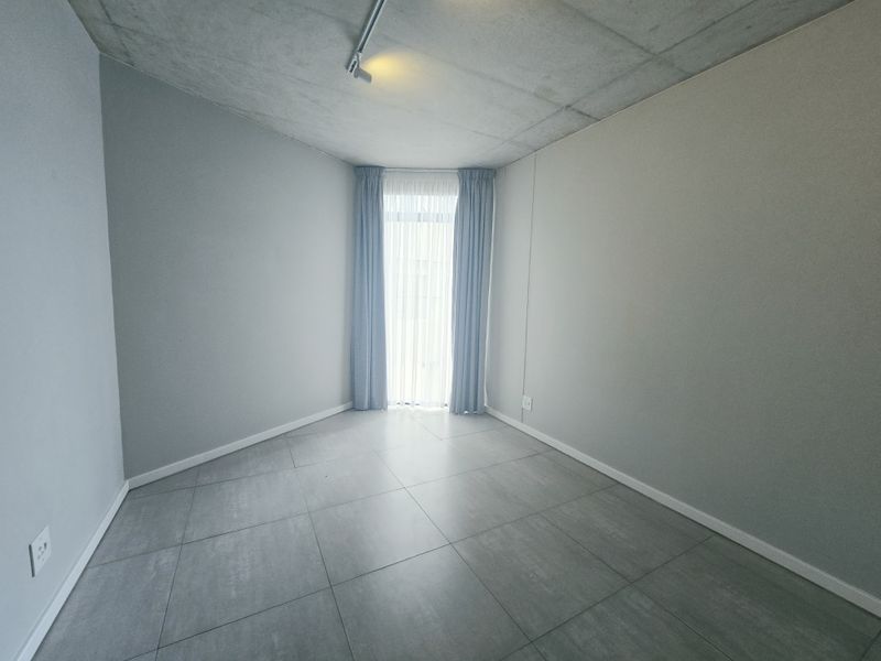 A clean, modern, and empty bedroom featuring an exposed concrete ceiling, large format floor tiles, and neutral wall finishes.