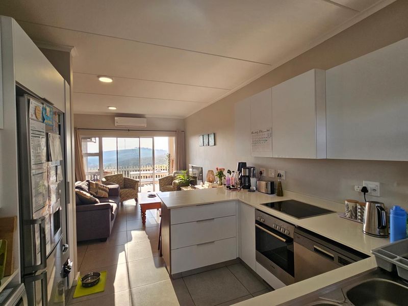 Modern open-plan kitchen and living area featuring sleek white cabinetry, integrated appliances, stone countertops, and sliding doors leading to a balcony with scenic views.