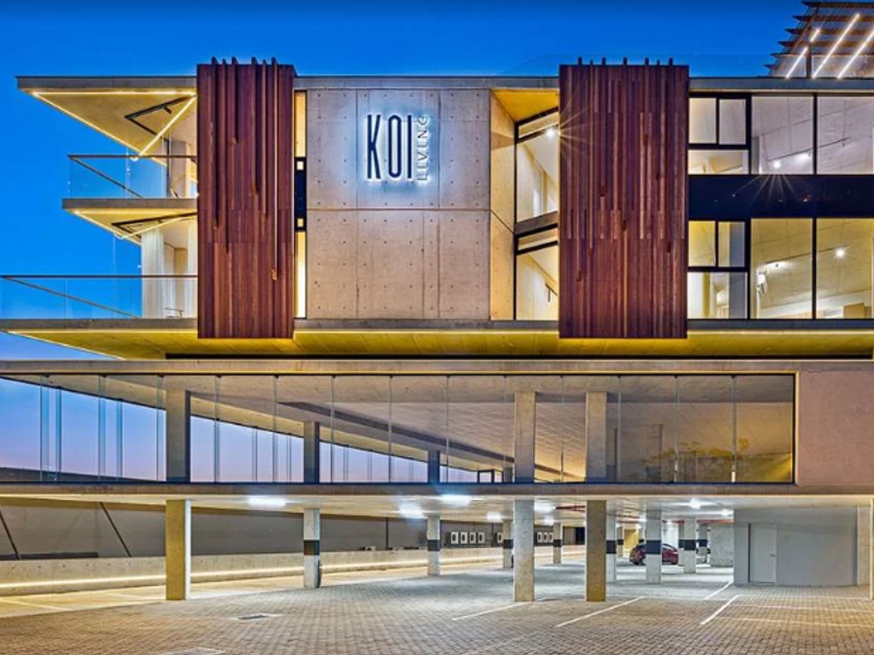 Modern apartment building facade featuring concrete architecture, timber accents, glass balconies, and a spacious covered ground-level parking area.
