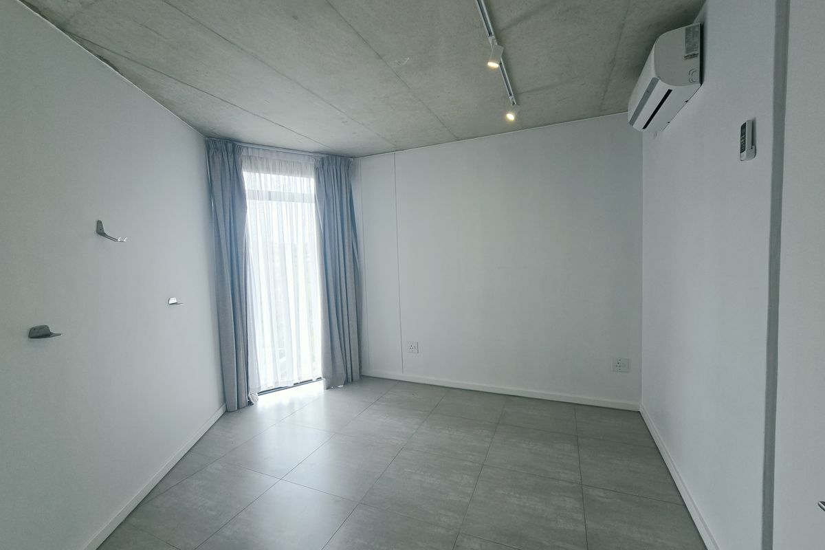 A bright, modern bedroom featuring an exposed concrete ceiling, tiled flooring, track lighting, and a split-system air conditioner with access to a balcony.