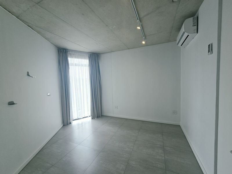 A bright, modern bedroom featuring an exposed concrete ceiling, tiled flooring, track lighting, and a split-system air conditioner with access to a balcony.