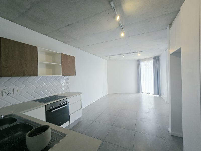Modern open-plan kitchen and living area featuring an exposed concrete ceiling, sleek cabinetry, herringbone backsplash, and large format floor tiles.
