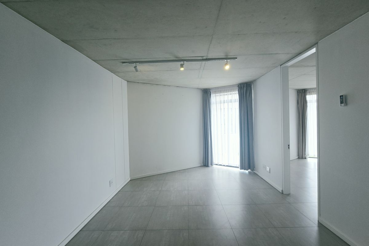 A bright, modern living area featuring an exposed concrete ceiling, sleek tiled flooring, and minimalist built-in cabinetry with track lighting.