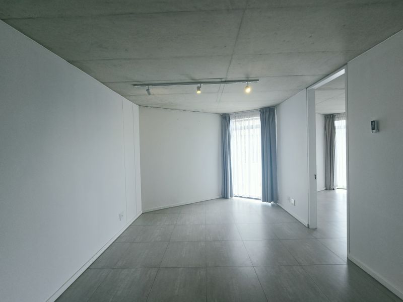 A bright, modern living area featuring an exposed concrete ceiling, sleek tiled flooring, and minimalist built-in cabinetry with track lighting.