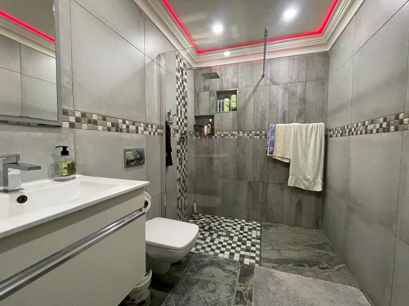 Modern bathroom featuring grey tiled walls, a wall-hung vanity, a walk-in shower with a rain shower head, and integrated LED ceiling lighting.