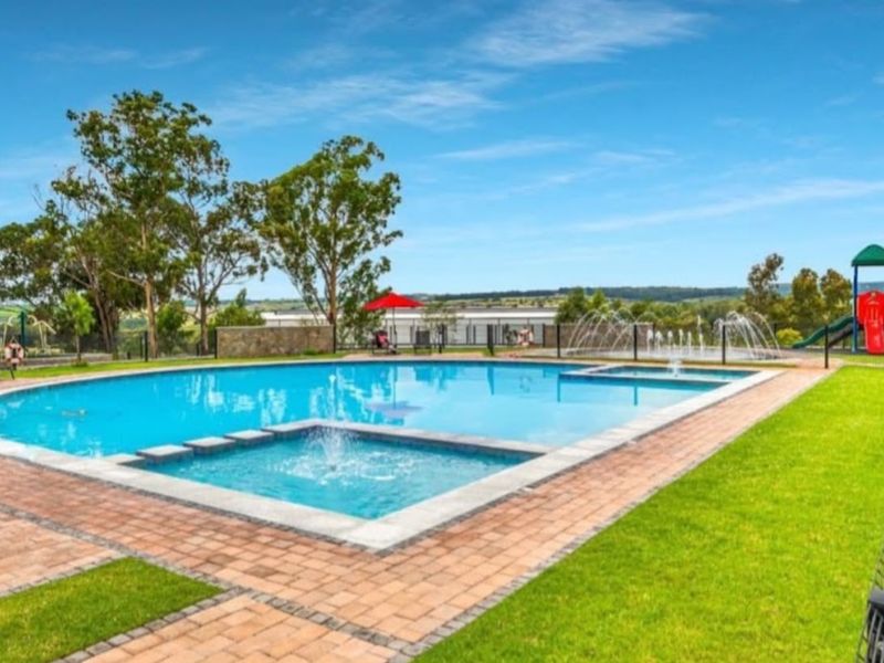 A well-maintained outdoor swimming pool area featuring a water fountain, paved surround, and landscaped lawn, set within a secure fenced perimeter.