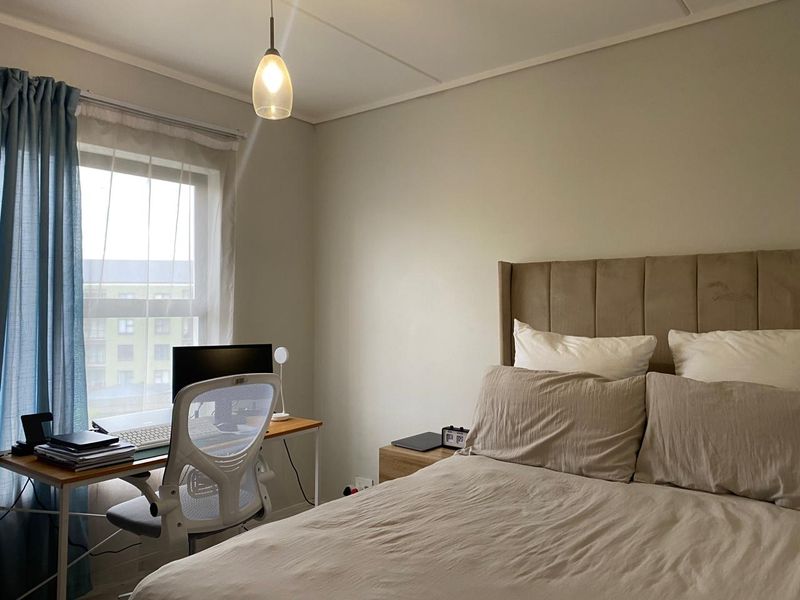 A well-lit bedroom featuring neutral wall tones, a window with natural light, and a functional layout suitable for a sleeping and workspace area.