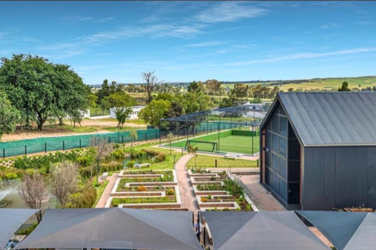 Aerial view of a well-maintained estate featuring landscaped gardens, a pond, and a modern sports facility with a metal-roofed building.