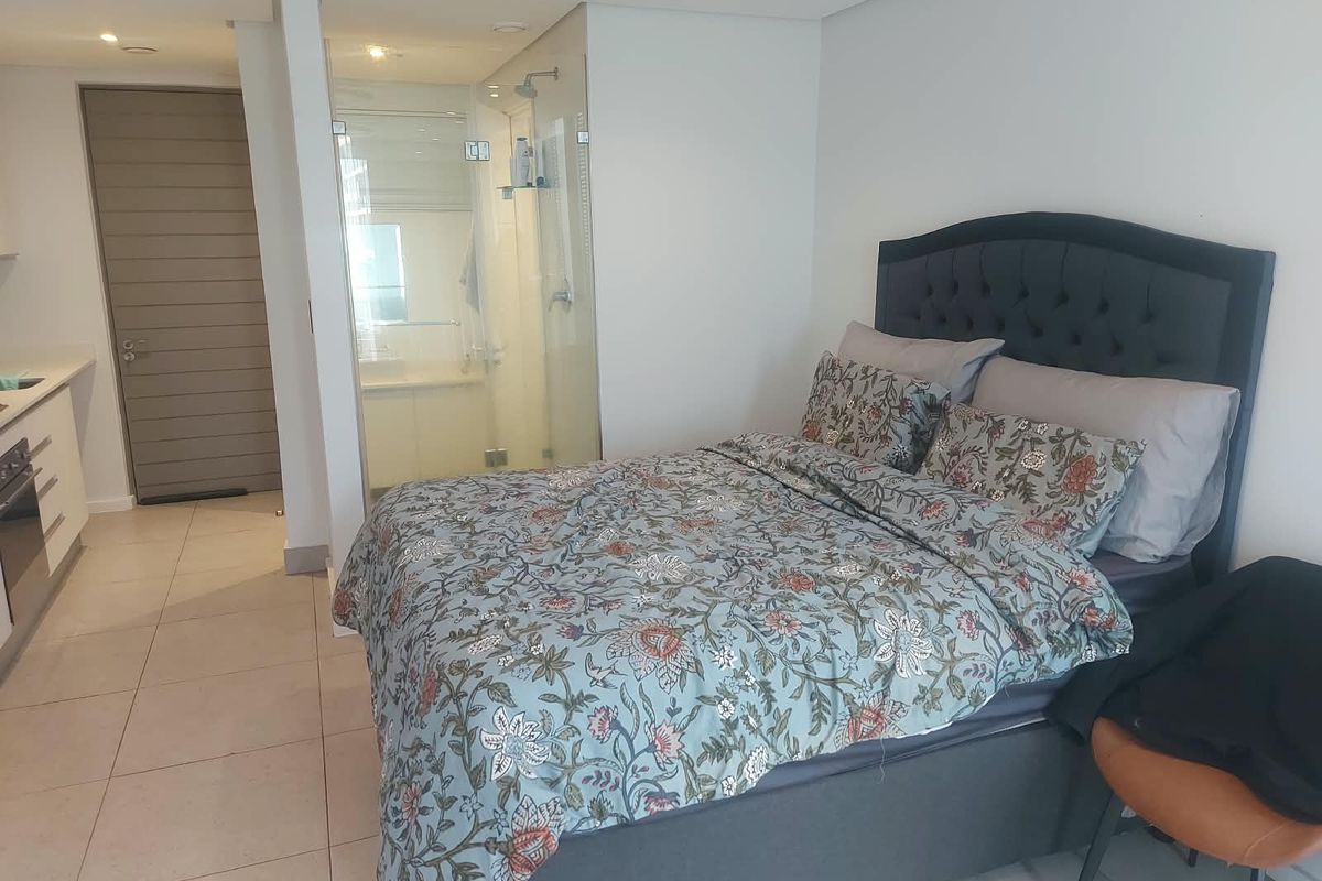 A modern bedroom featuring an integrated en-suite bathroom with a glass shower enclosure, tiled flooring, and recessed lighting in a contemporary apartment setting.