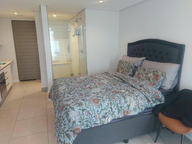 A modern bedroom featuring an integrated en-suite bathroom with a glass shower enclosure, tiled flooring, and recessed lighting in a contemporary apartment setting.