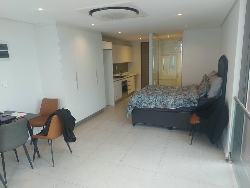 Modern open-plan studio apartment featuring a compact kitchen with built-in appliances, tiled flooring, recessed lighting, and an en-suite glass-enclosed bathroom.
