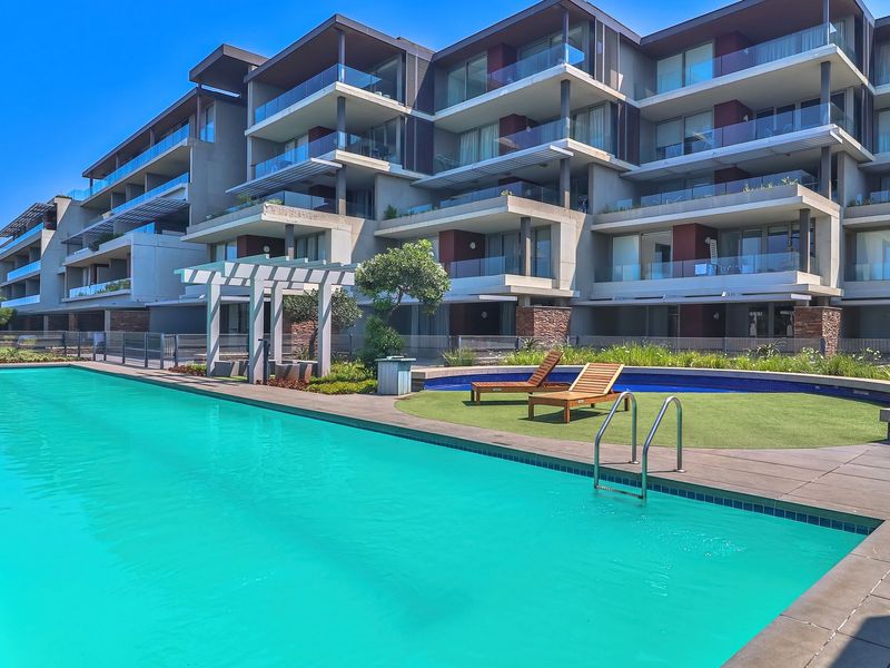 A well-maintained communal swimming pool area featuring a tiled deck, pergola, and modern apartment building facade with glass balconies.