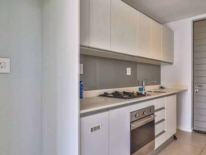 A modern, compact kitchen featuring sleek white cabinetry, stone countertops, a gas hob, built-in oven, and a neutral glass splashback.