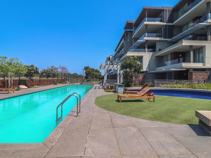 A well-maintained communal swimming pool area featuring a tiled deck, artificial lawn, and modern apartment building architecture in the background.