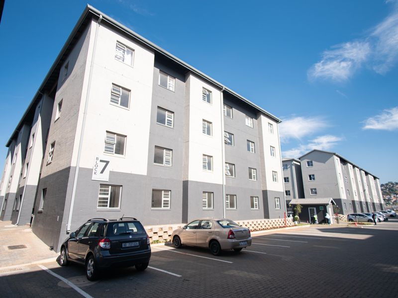A view of a multi-story apartment block featuring a modern painted facade, casement windows, and an adjacent paved parking area.