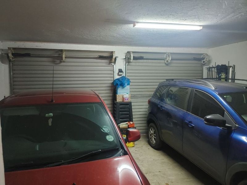 A functional double garage featuring two roller doors, concrete flooring, and overhead fluorescent lighting, providing secure parking space.