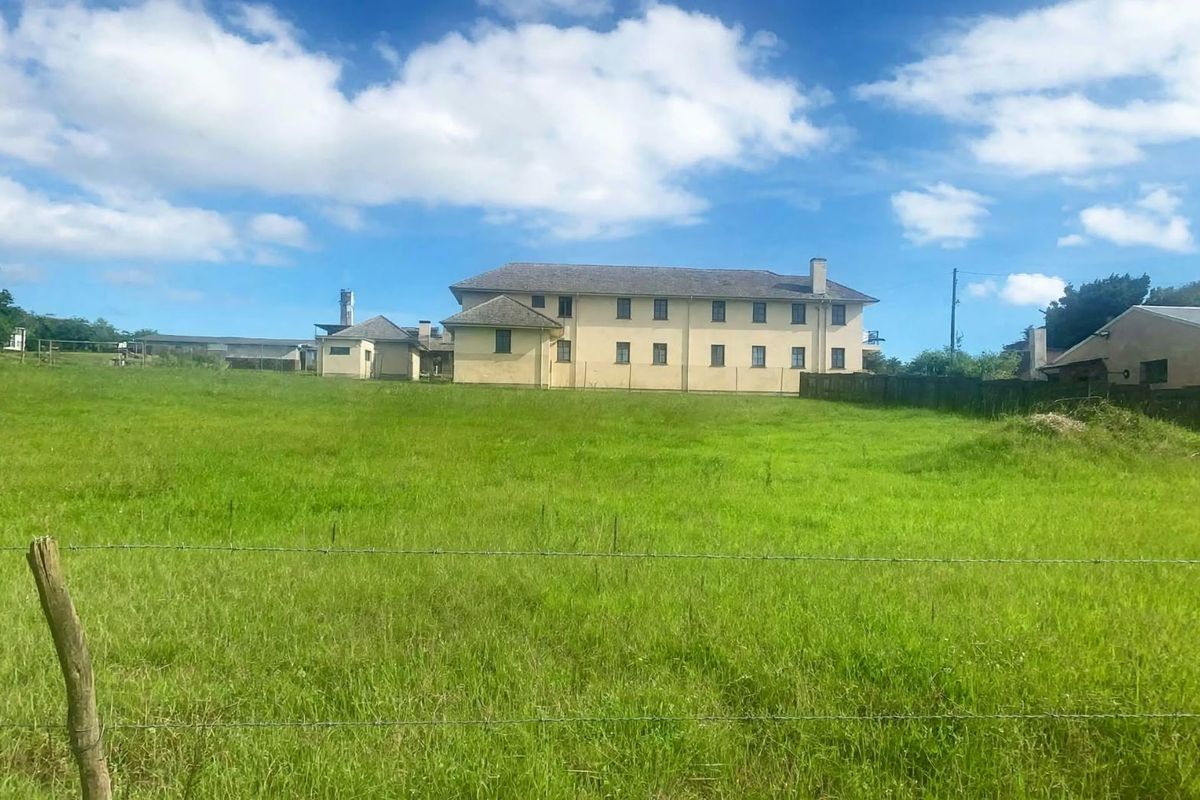 A view of the rear elevation of a large residential building set within a spacious, grassy field enclosed by a wire fence.