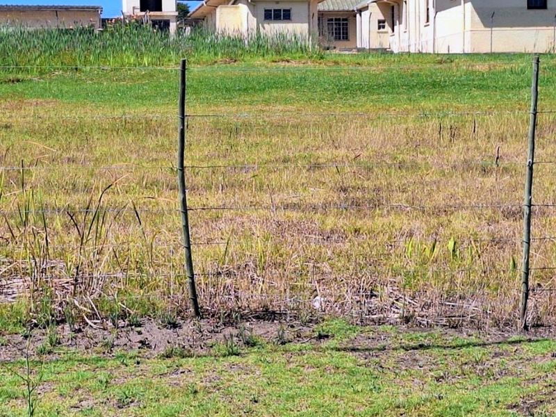 A view of a vacant residential plot featuring wire fencing and wooden posts, with neighboring buildings visible in the background.