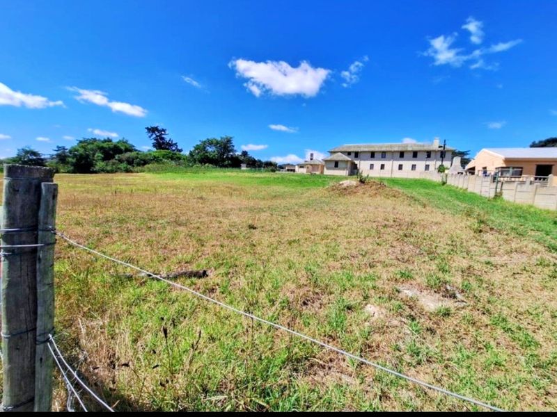 A vacant residential plot featuring a wire fence boundary, open grassed terrain, and views of neighboring properties under a clear blue sky.