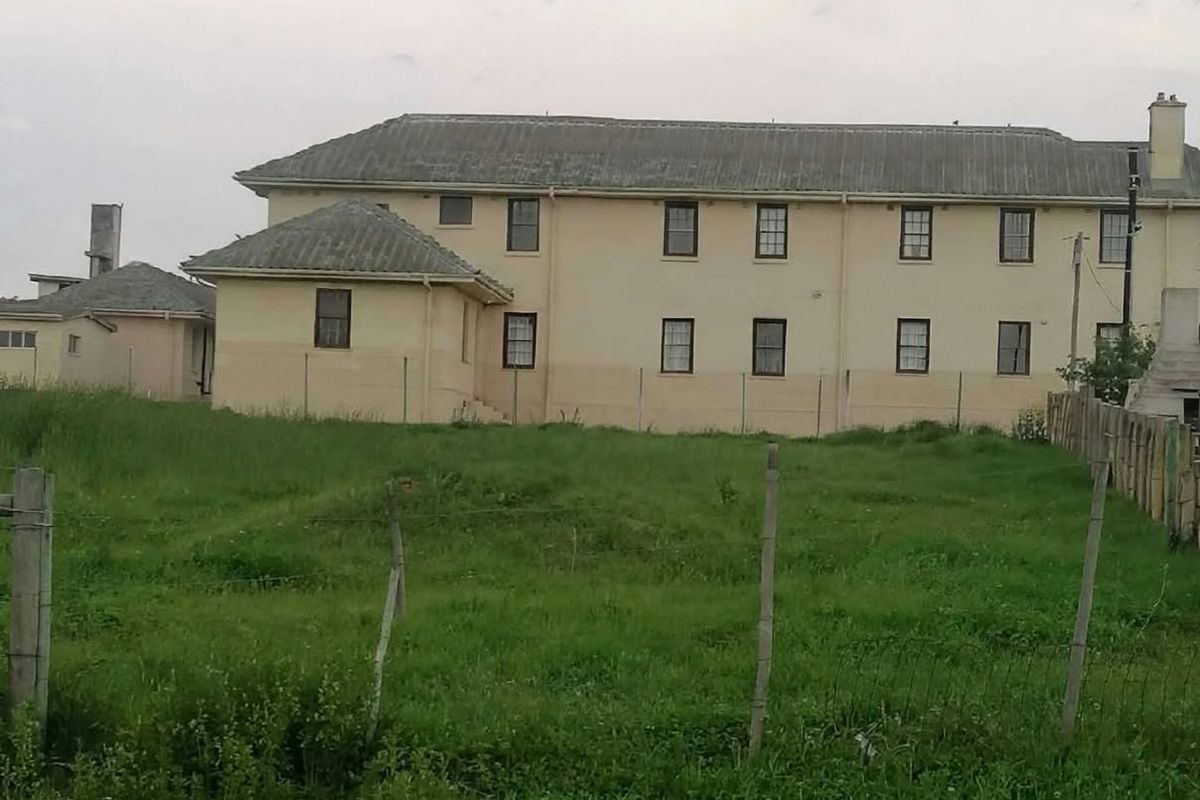 Rear view of a multi-story residential building featuring a pitched roof, chimney, and surrounding wire fencing on a grassy plot.