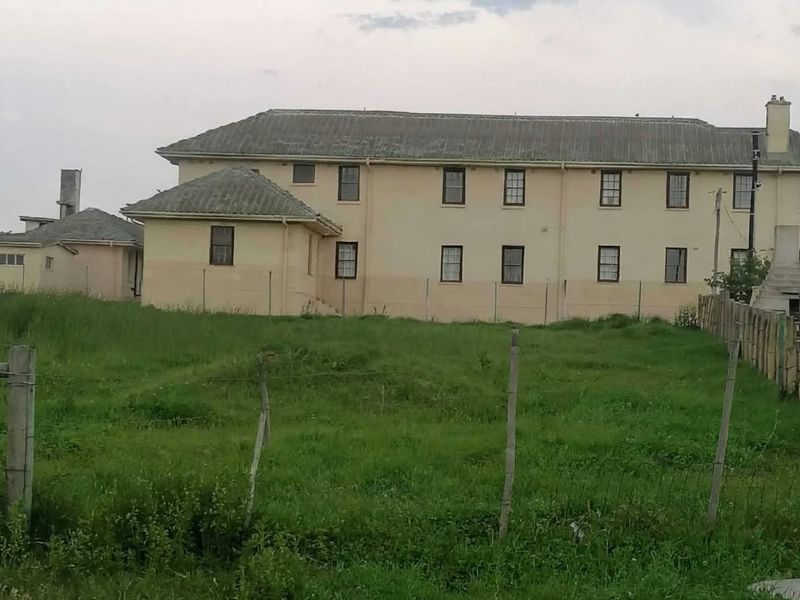 Rear view of a multi-story residential building featuring a pitched roof, chimney, and surrounding wire fencing on a grassy plot.
