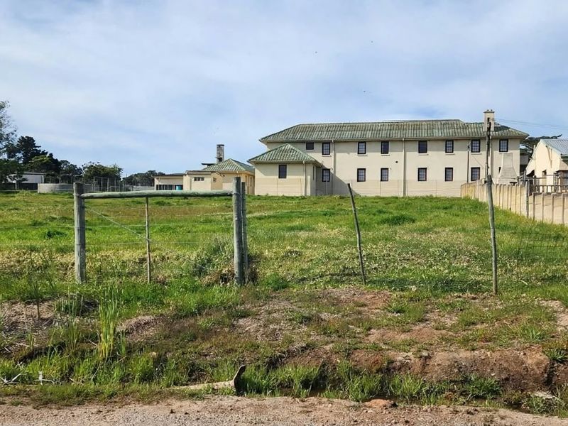 A view of a vacant residential plot featuring grass cover and wire perimeter fencing, with neighboring buildings visible in the background.