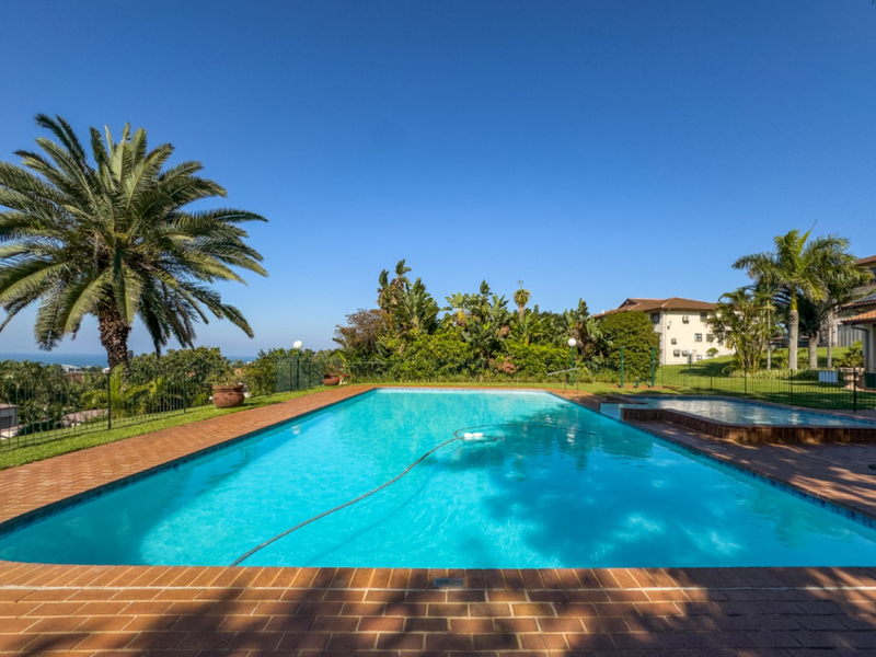 A well-maintained outdoor swimming pool area featuring brick paving, perimeter fencing, and established garden landscaping with scenic views.