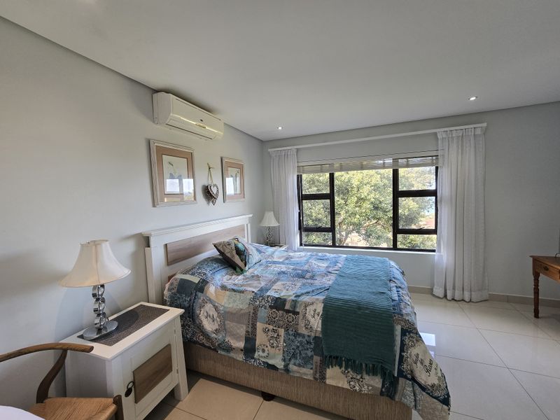 A well-maintained bedroom featuring tiled flooring, recessed downlighting, and a wall-mounted split-system air conditioner, with large windows providing natural light and garden views.