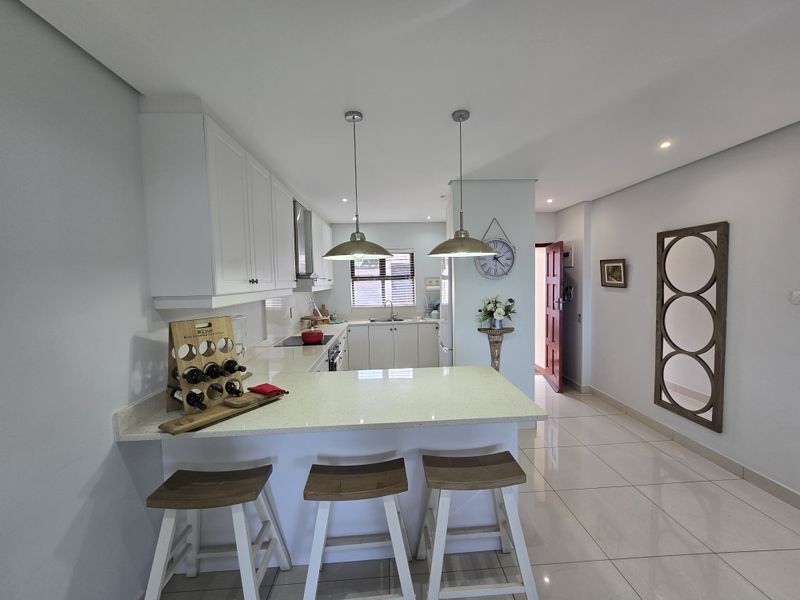 Modern open-plan kitchen featuring white cabinetry, quartz countertops, a breakfast bar, and sleek porcelain floor tiles, seamlessly flowing into the living area.